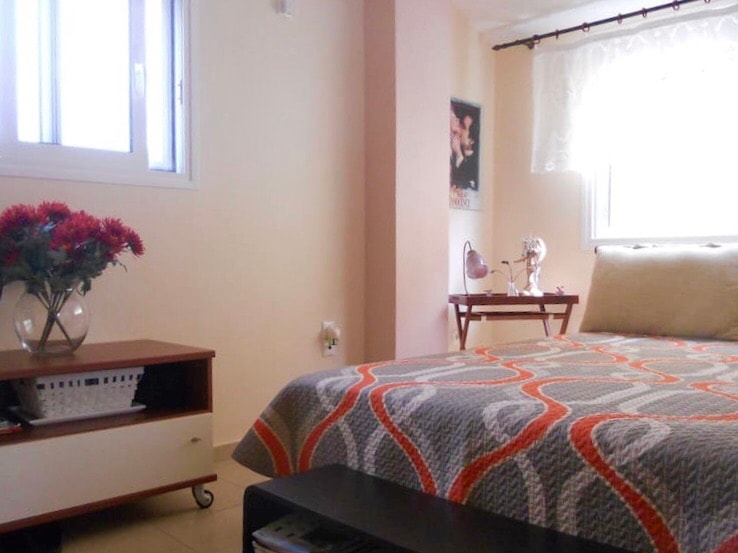 A well-lit bedroom features a spacious bed with a stylish patterned cover. A wooden nightstand holds a small lamp and a decorative vase with flowers. Natural light filters through the window, enhancing the calm ambiance.