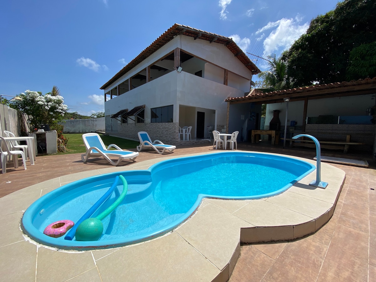 A refreshing pool in a unique shape is surrounded by a wooden deck. Sun loungers are positioned nearby, along with a table and chairs beneath a covered area. The house with a textured exterior showcases multiple upper-level windows, and greenery is visible in the background.