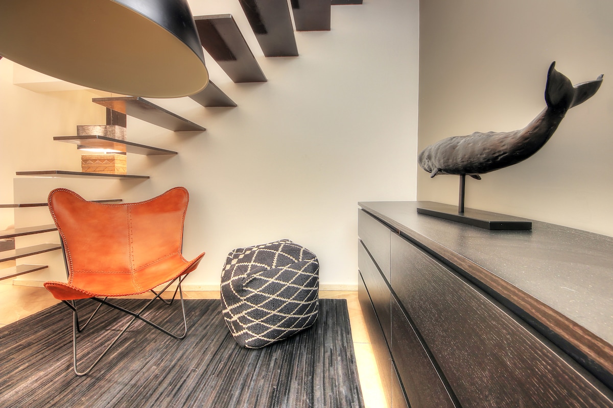 A modern interior showcases a leather butterfly chair paired with a textured floor pouf, situated on a striped rug. A sleek cabinet contrasts with the soft curves of the decorative whale sculpture, while a floating staircase leads to the upper level, enhancing the open space.