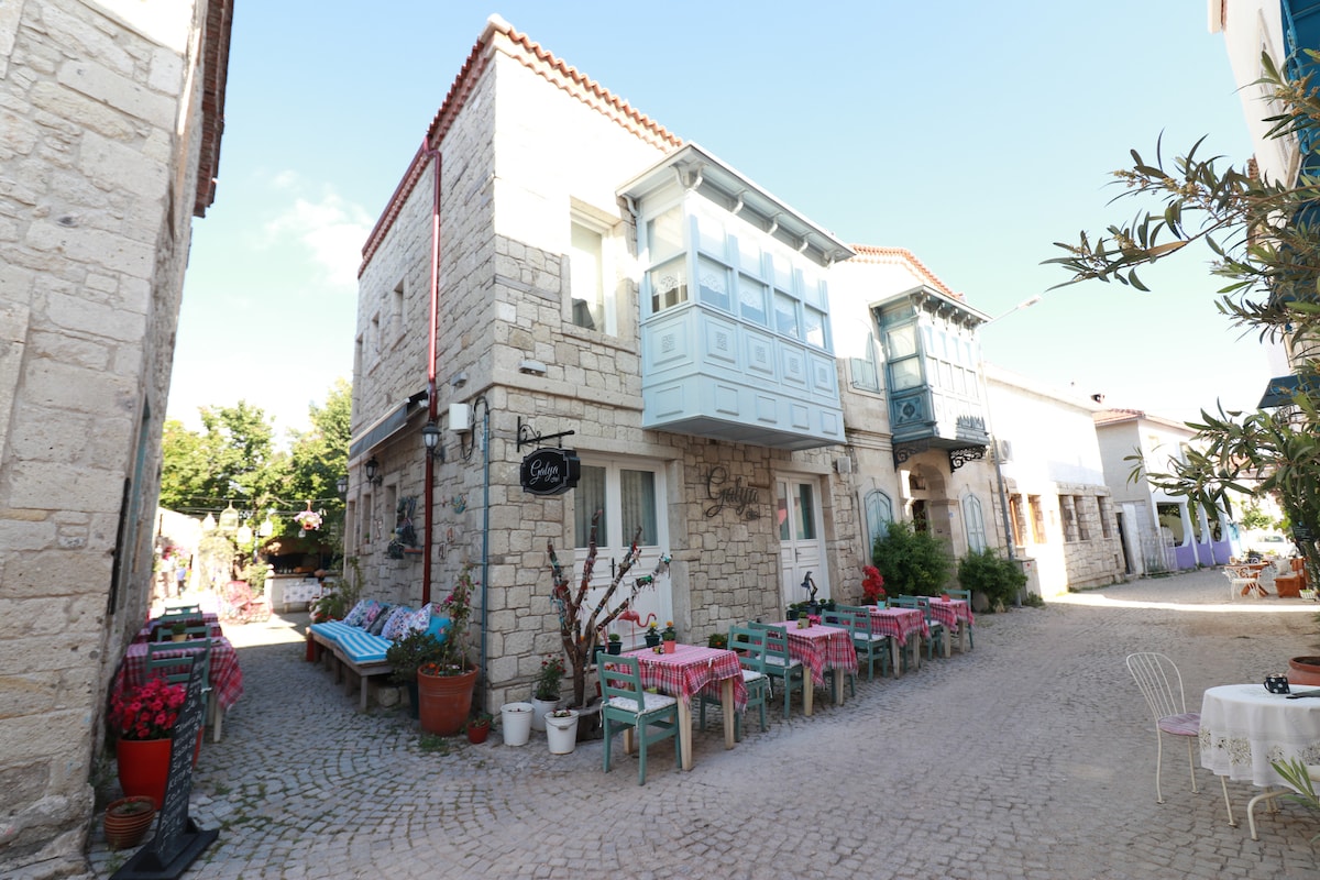 A charming street view showcases a stone building with a light blue bay window. Outdoor dining tables with colorful tablecloths are set along the cobblestone pathway, framed by pots of flowers, creating a welcoming environment in a pedestrian-friendly area.
