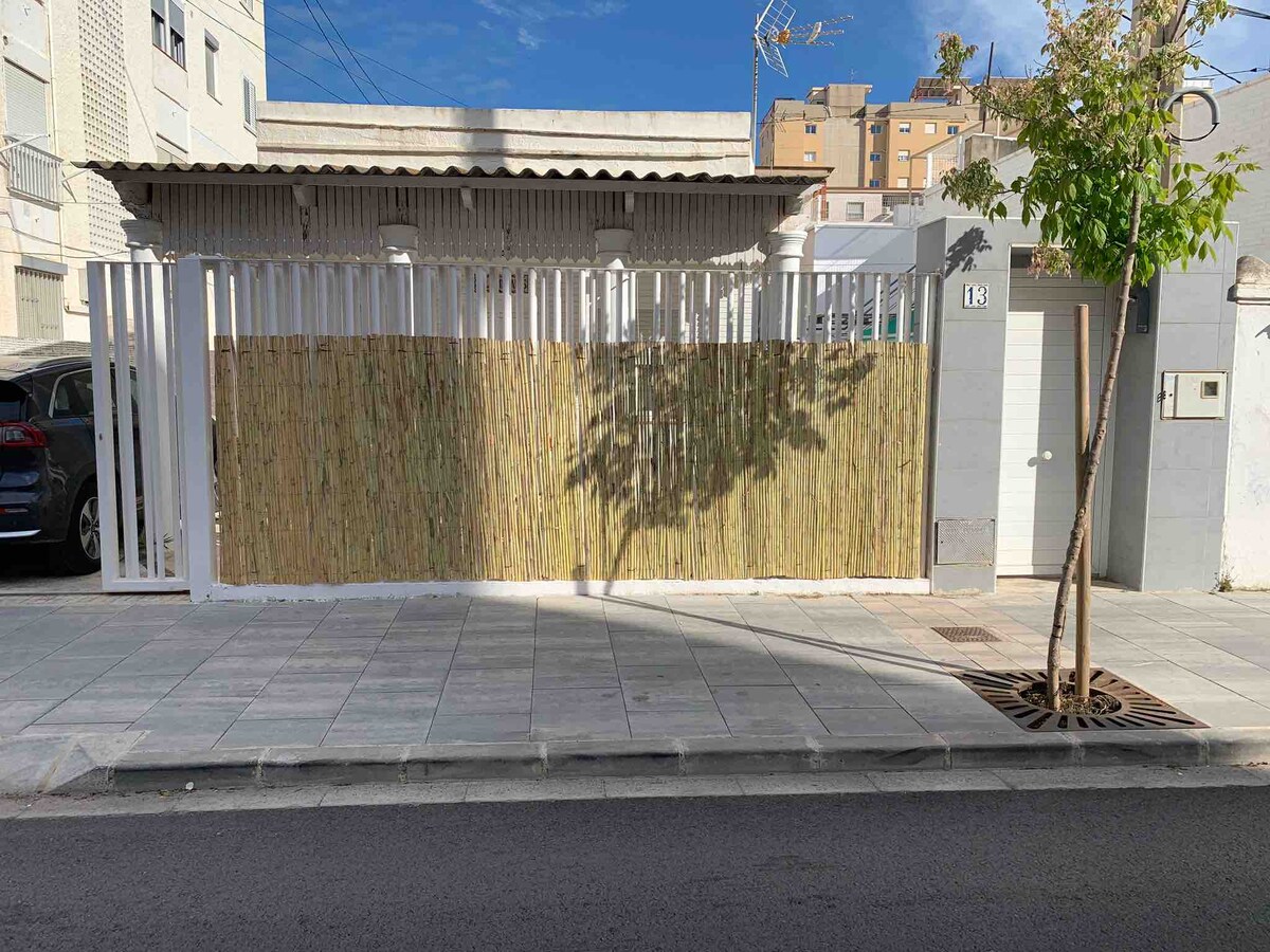 The exterior of the property is showcased with a bamboo fence providing privacy. A tree is planted near the entrance, and the facade features a light-colored wall. A sidewalk runs along the front, with a glimpse of residential buildings in the background.