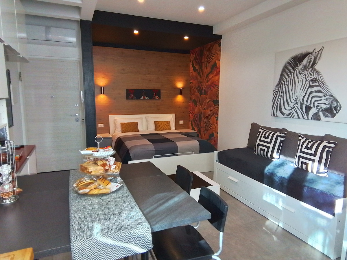 An open space features a cozy sleeping area with a spacious bed framed by decorative wall art. A stylish sofa bed is positioned adjacent to a modern dining table, accompanied by black chairs. The kitchen area is equipped with contemporary appliances, enhancing functionality.