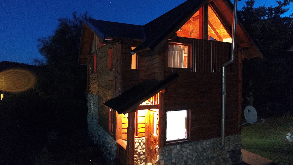 The exterior of a spacious wooden cabin is illuminated at dusk, showcasing a welcoming ambiance. Large windows allow warm light to spill out, revealing a blend of natural materials and a stone foundation. Surrounding greenery complements the tranquil setting.