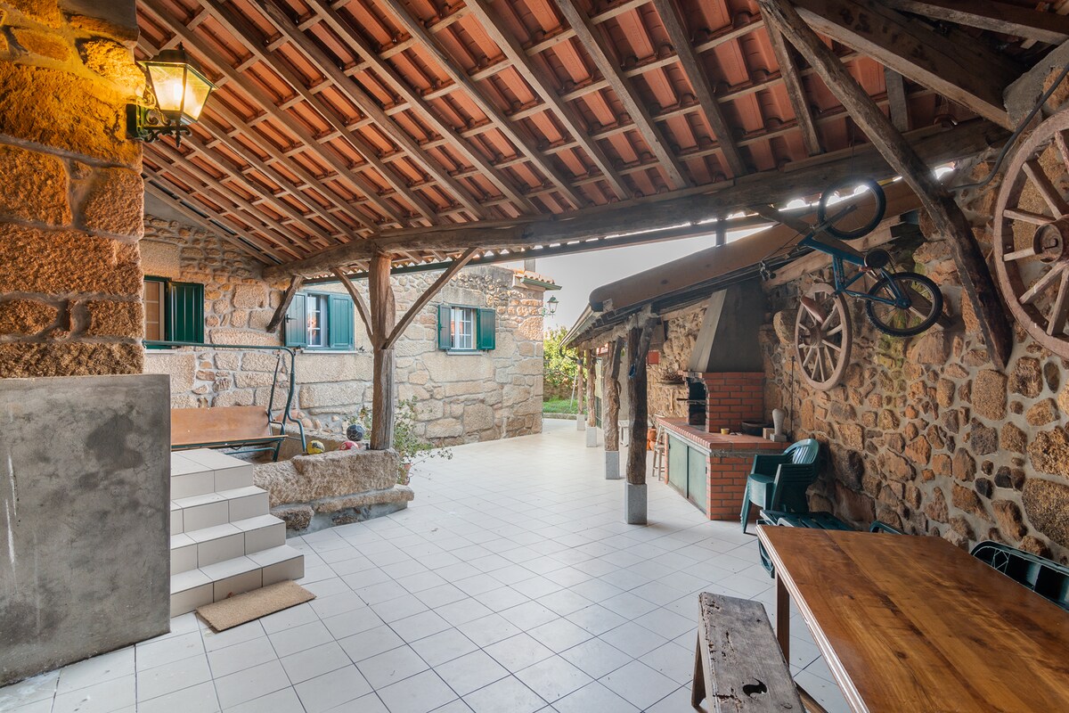 An outdoor space is presented, featuring a stone structure with extensive tiled flooring and rustic wooden beams overhead. An outdoor kitchen area includes a grill and work surfaces. A dining table is situated to one side, while nature views are framed by green window shutters.