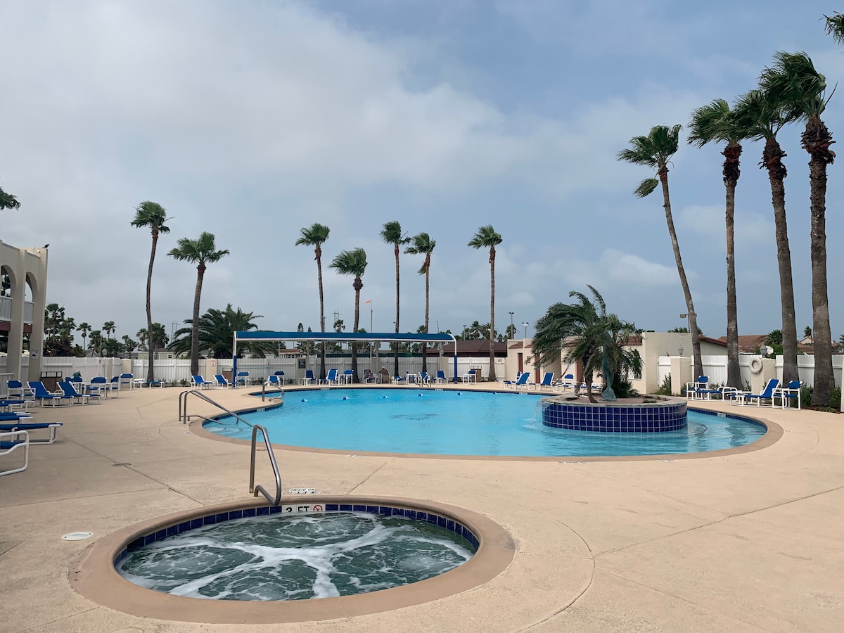 The resort pool area is surrounded by palm trees and features a large swimming pool alongside a connected hot tub. Sun loungers are arranged around the pool deck, offering ample space for relaxation. The sky appears partly cloudy, providing gentle shade.