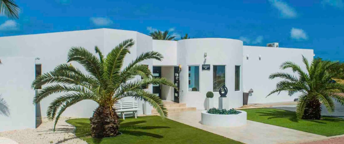 A modern villa features smooth white walls and large windows framed by palm trees. A sculpture is displayed in the front yard, which is complemented by well-maintained grass and decorative plants. A seating area is positioned beside the entrance, offering an inviting outdoor experience.