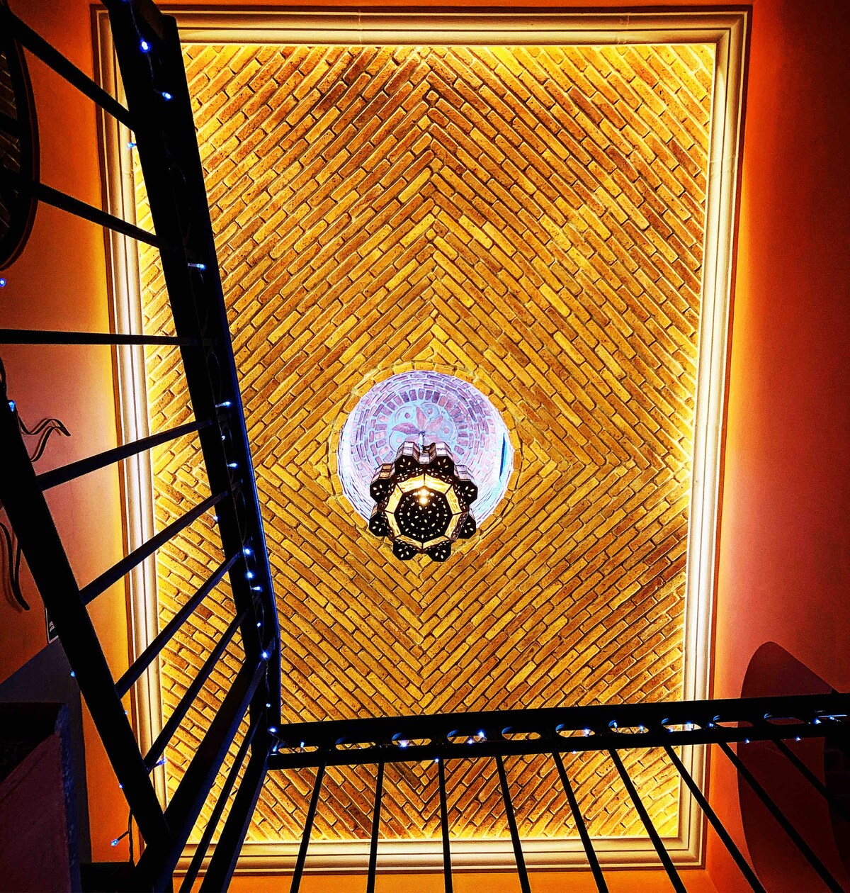 The view captures the intricately designed ceiling adorned with a decorative light fixture. The brick pattern is accentuated by warm lighting, enhancing the architectural details. A black metal staircase frames the image, leading up from the lower level.