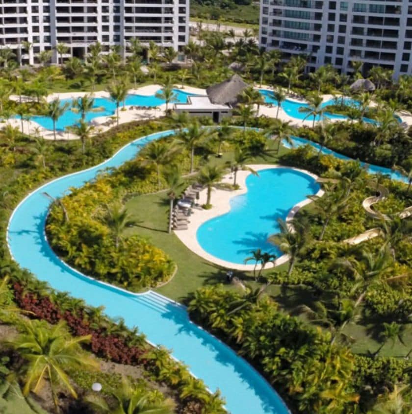 Aerial view of a resort-style pool area featuring meandering water channels surrounded by lush greenery. Multiple pools with varying shapes are interspersed among palm trees and vibrant landscaping, providing a serene and inviting atmosphere for relaxation.