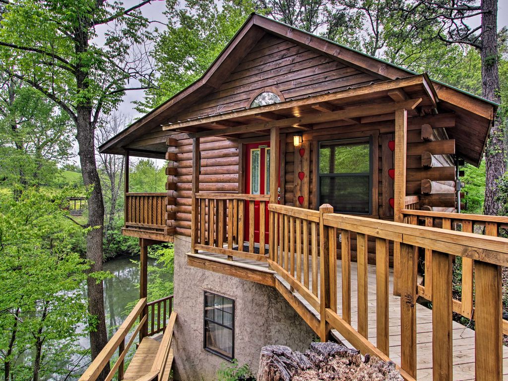 A cabin exterior is showcased, featuring a rustic wooden structure with a covered porch and bright red door. Surrounding greenery and trees provide a natural setting, while the cabin is elevated above the ground, with a wooden walkway leading to the entrance.
