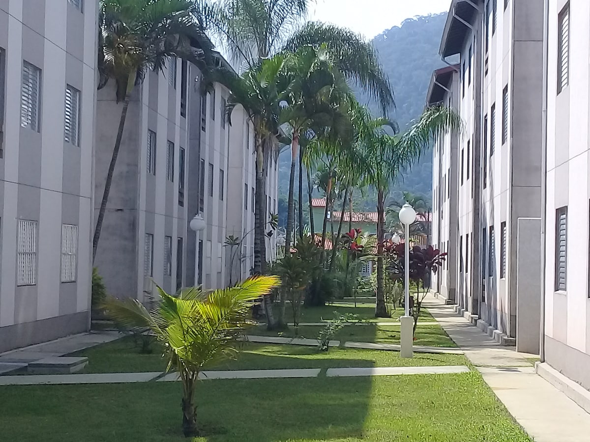 A serene courtyard is presented, featuring well-maintained grass and lined by palm trees. Sunlight filters through the leaves, casting soft shadows on the pathways that connect the buildings. The surrounding mountains create a tranquil backdrop, enhancing the feeling of openness.