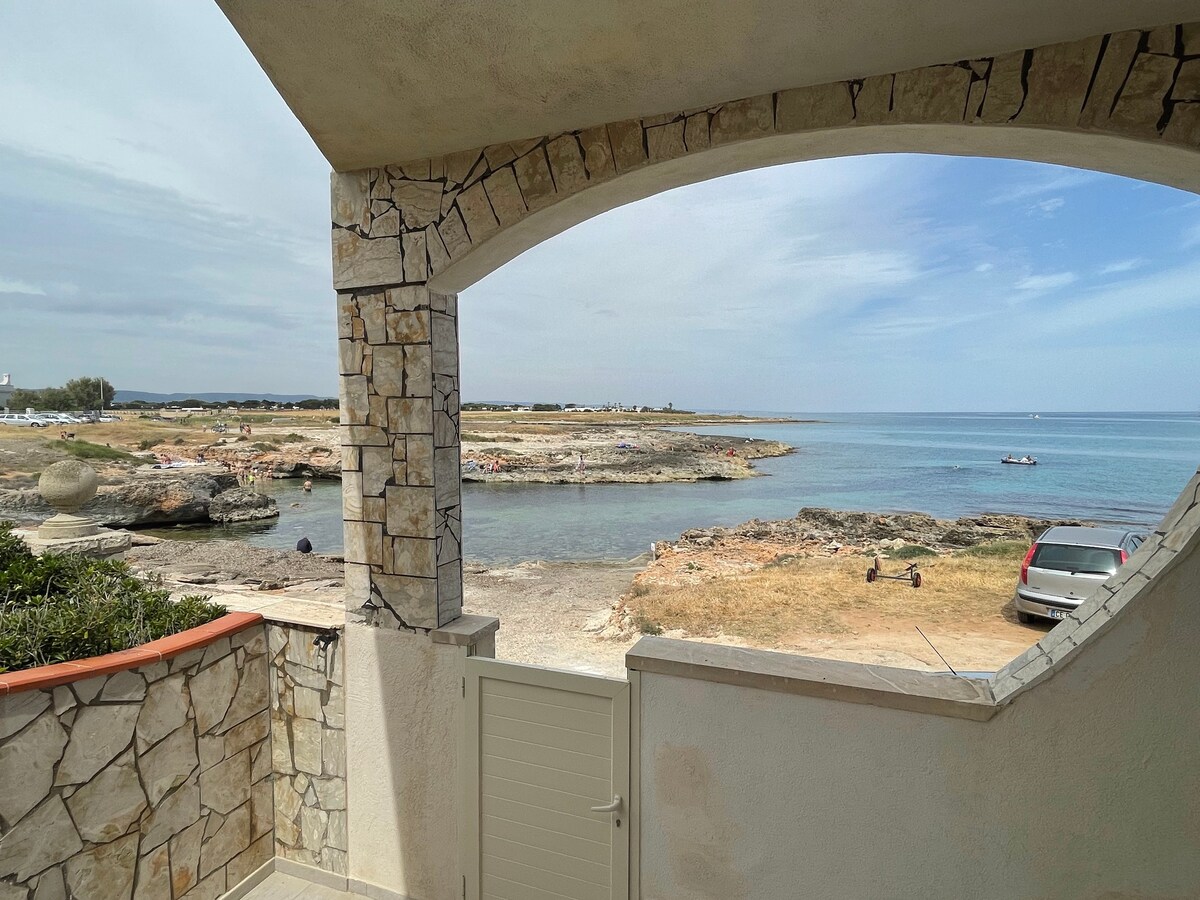 A stone archway frames a serene coastal view, revealing calm waters and rocky shores. The landscape features patches of greenery, with a vehicle parked nearby. The horizon blends seamlessly with the sky, creating a peaceful atmosphere just steps from the property.