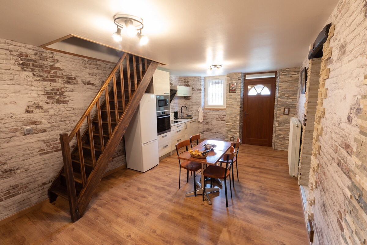 A warm and inviting loft space features a wooden staircase leading to the upper level. The kitchen area, equipped with modern appliances, is complemented by a dining table surrounded by chairs. Exposed brick walls and wood flooring enhance the cozy aesthetic.