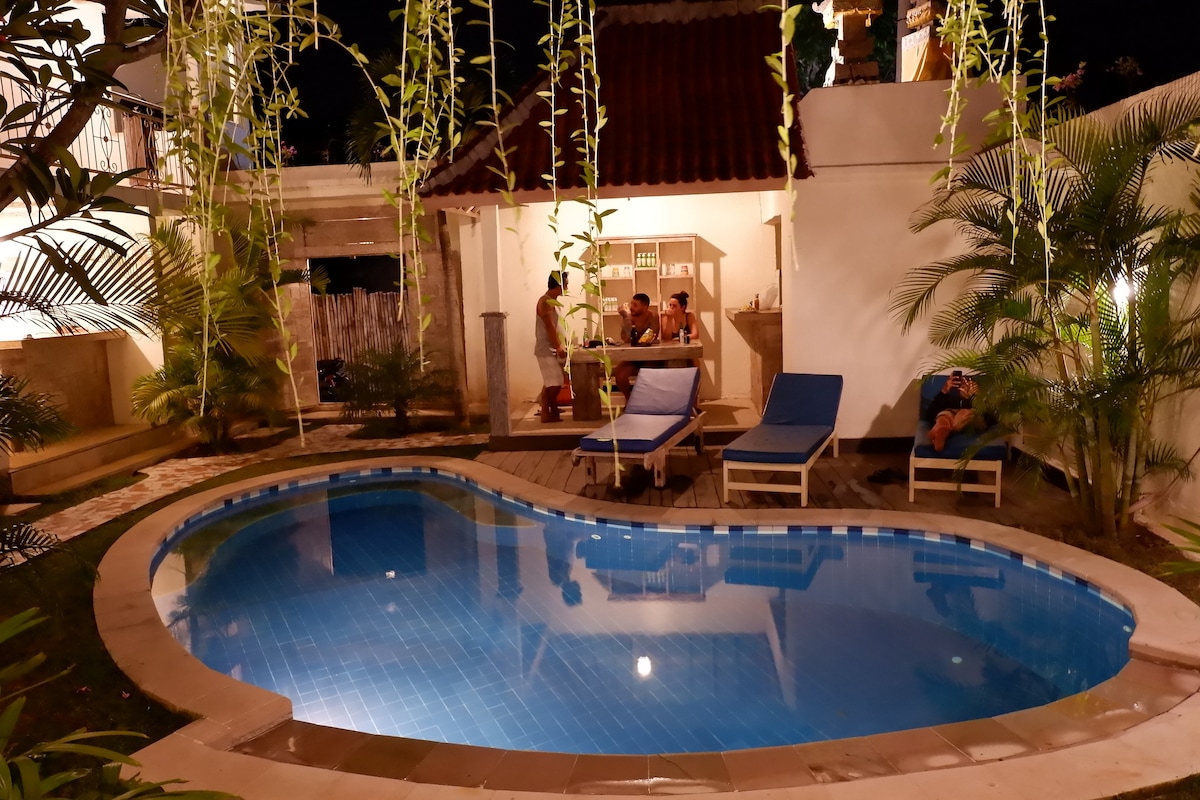 A serene swimming pool is framed by lush tropical plants, with a warm light illuminating the area. Comfortable lounge chairs are positioned nearby, inviting relaxation. A small seating area can be seen under a shaded structure, providing a cozy spot for gatherings.