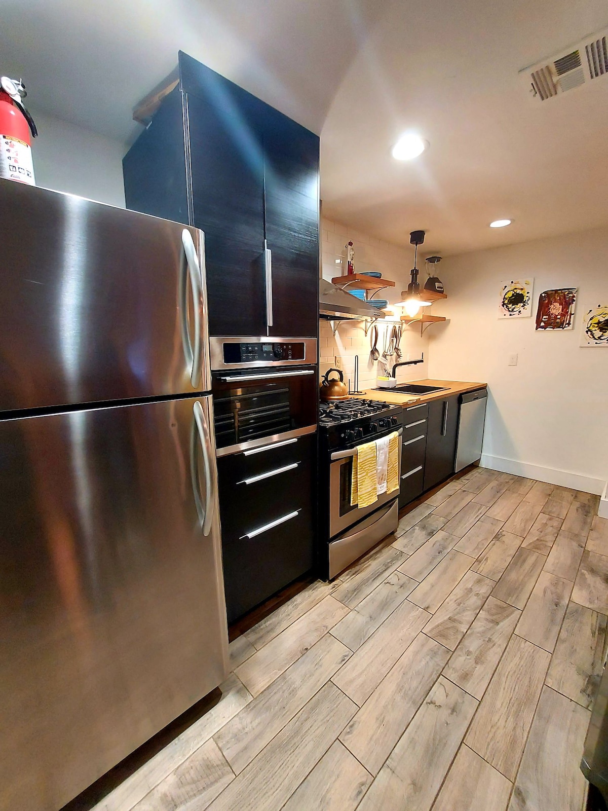A modern kitchen features sleek stainless steel appliances, including a refrigerator and oven. Wood countertops provide a warm contrast, and various kitchen utensils are organized on the shelves above. The tile flooring contributes to the contemporary design, illuminated by overhead lighting.