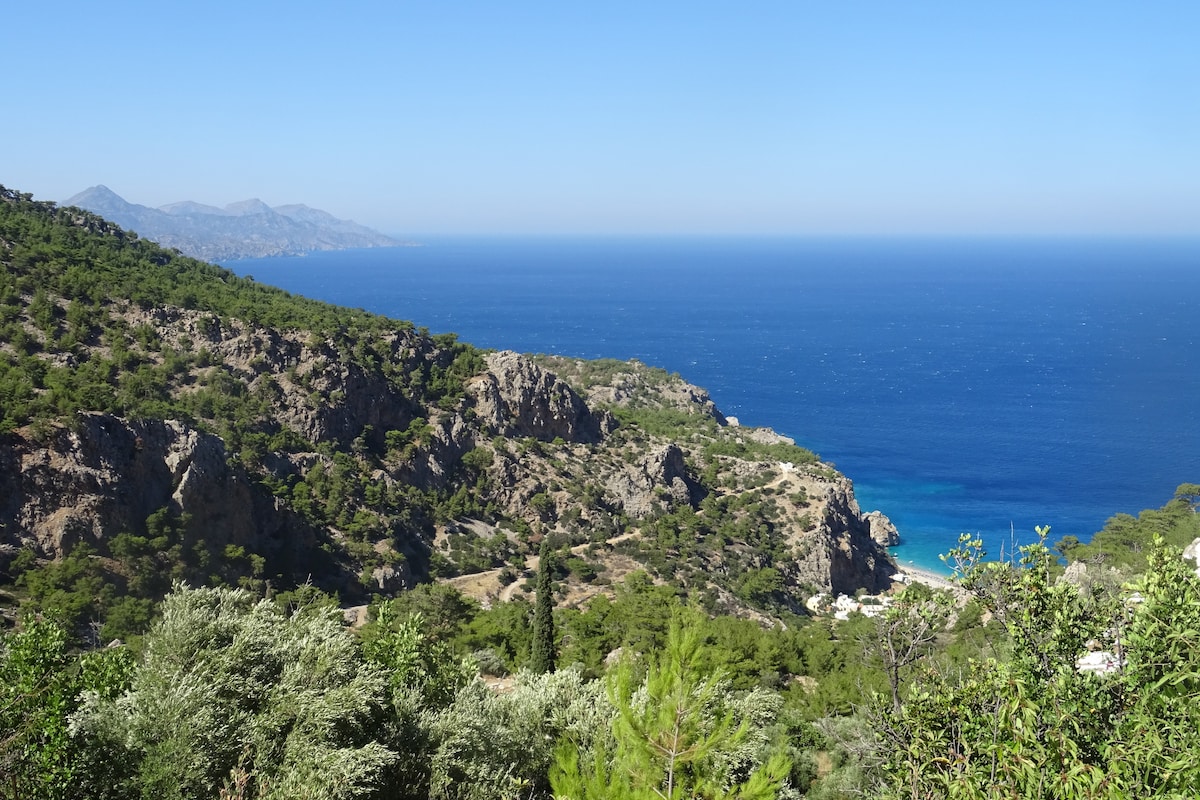 A panoramic view of a rugged coastline is captured, showcasing lush green hillsides that descend towards the azure sea. The serene beach is visible in the distance, framed by rocky cliffs and dotted with trees, highlighting the natural beauty of the island's landscape.