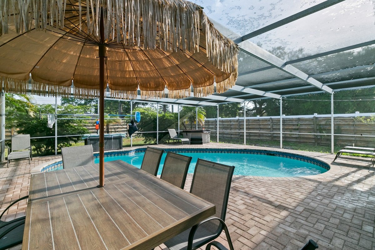 An inviting outdoor space showcases a private pool surrounded by a spacious patio. A thatched umbrella provides shade over a dining table with multiple chairs. The pool's blue water is visible, framed by a fenced yard and lush greenery in the background.