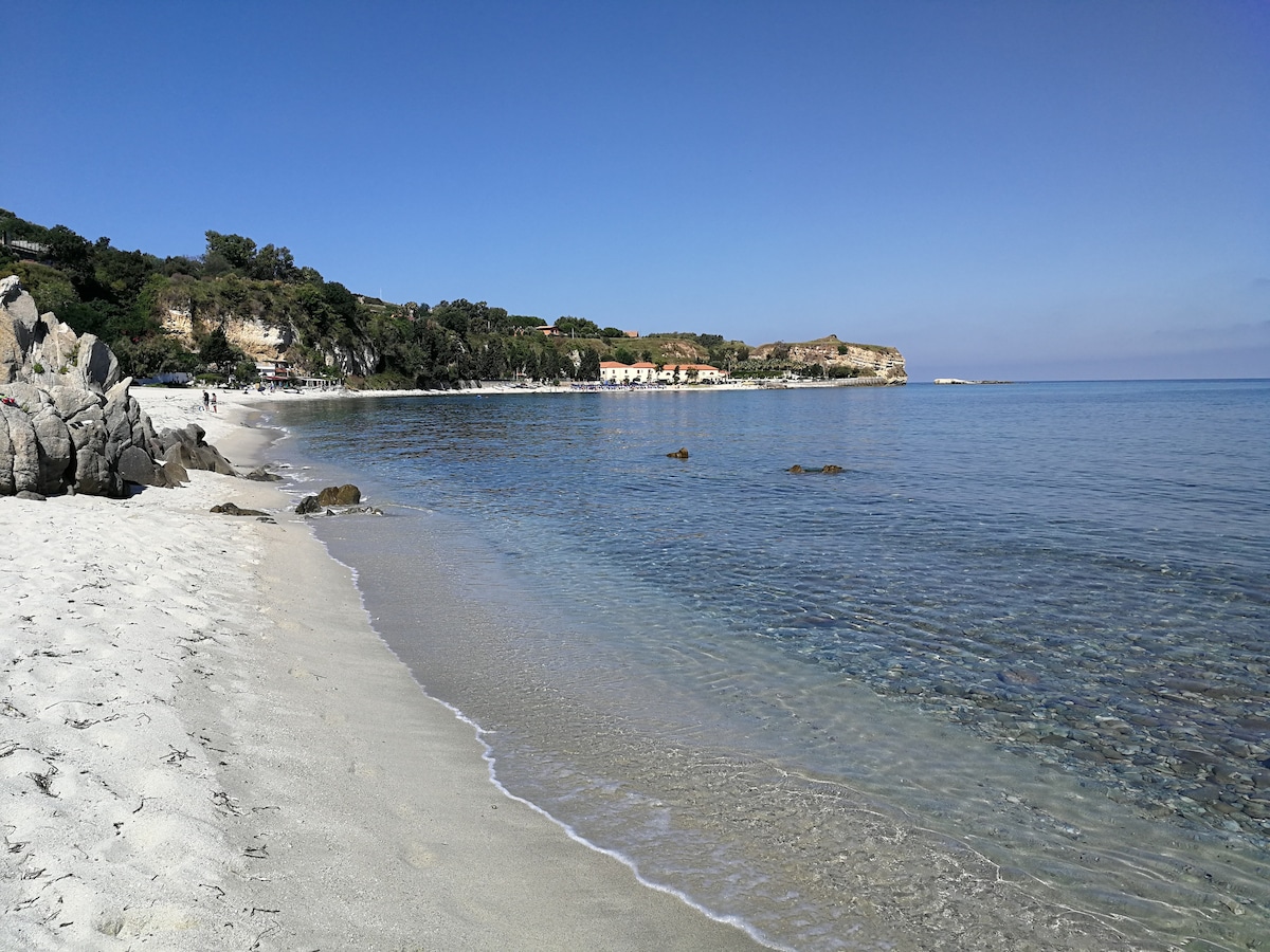 A serene beach scene showcases a gentle curve of soft sand leading to clear, calm waters. The scenery is complemented by lush greenery and rocky formations in the background, creating a tranquil coastal setting.