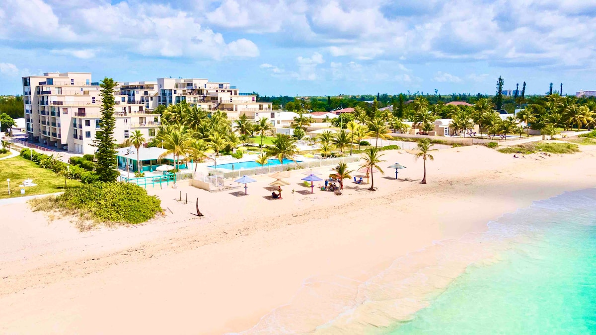 The image showcases a spacious beachfront view of the Coral Beach Hotel, featuring modern architecture surrounded by lush palm trees. A swimming pool is visible near the hotel, along with beach chairs and umbrellas set on soft, sandy beach, leading to vibrant turquoise waters.