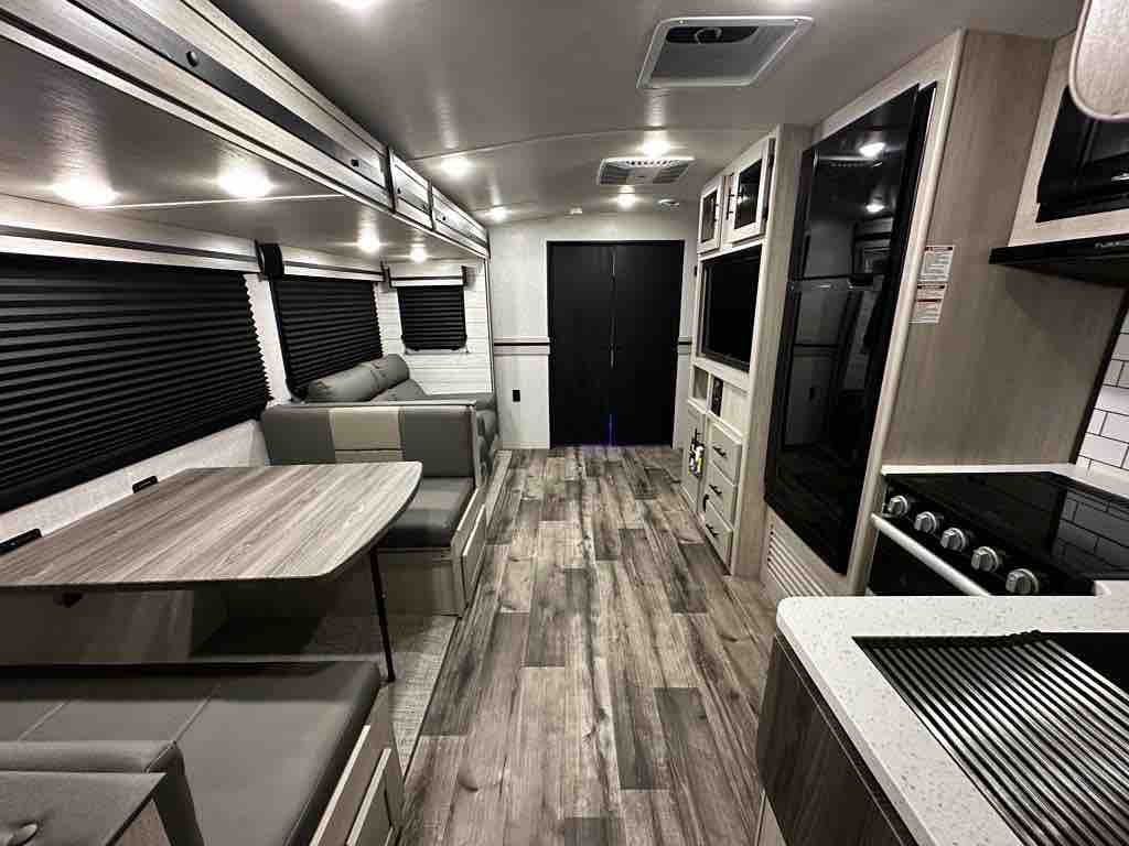 The interior of a modern RV is presented, featuring a spacious layout with light-colored cabinetry and dark blinds. A convertible dining table is visible alongside a sofa, while a kitchenette with a stovetop and sleek countertops is showcased. A dark door leads to another area.