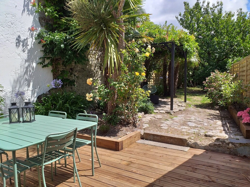 The outdoor terrace features a wooden deck with a dining table and chairs surrounded by lush greenery. A paved pathway leads towards a garden area enriched with flowering plants and trees, including palm and rose bushes. A pergola provides shaded seating.