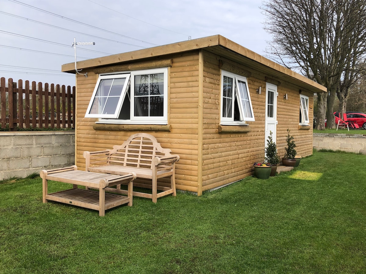 A charming cabin is presented, featuring a sloped roof and wooden exterior. Two large windows allow natural light to enter, while a bench and coffee table are positioned on the grassy area in front. Potted plants enhance the outdoor space, creating a welcoming atmosphere.