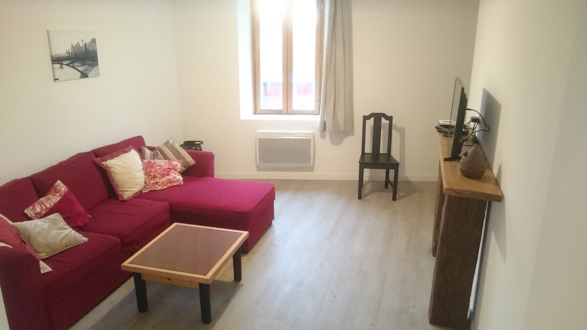 A modern living area features a red L-shaped couch adorned with decorative cushions. A wooden coffee table occupies the center, and a flat-screen TV is mounted on the wall. Natural light enters through a large window with light curtains, illuminating the neutral-toned flooring.