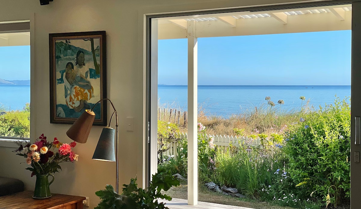 A view of the serene ocean is framed by glass doors, inviting natural light into the space. A wooden table holds a lamp and a vase of flowers. A colorful painting hangs on the wall, adding an artistic touch, while lush greenery is visible outside.