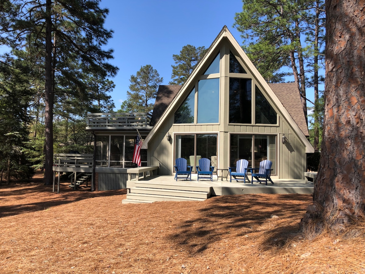 The exterior of a modern A-Frame house is shown, featuring large windows that allow natural light to fill the space. A welcoming deck with several blue chairs provides an inviting outdoor seating area. The surroundings are lined with tall pine trees and a bed of pine needles.