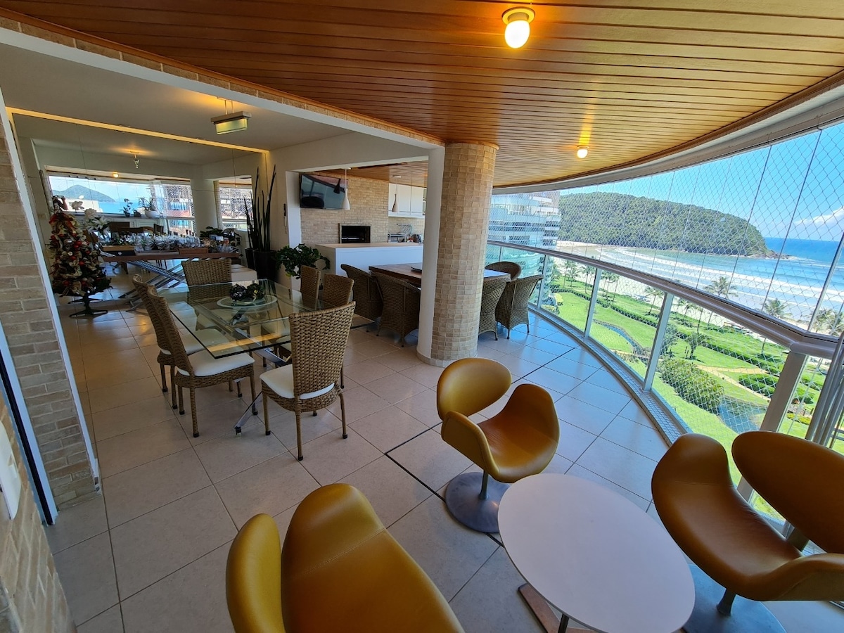 A spacious balcony with wooden ceiling features a dining area and comfortable seating. Large glass doors provide a panoramic view of the beach and surrounding landscape. A small coffee table sits at the center, complementing the modern design of the wicker furniture.