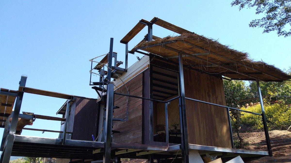 A wooden structure elevated on stilts is featured, showcasing a distinct rustic design with a thatched roof. Surrounding greenery enhances the natural setting, and a clear blue sky is visible overhead, providing a bright and open atmosphere.