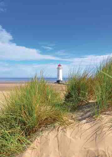 A coastal view features a white lighthouse with a red top, standing prominently against a clear blue sky. Grassy dunes frame the foreground, leading to a sandy beach that extends toward the horizon, where the sea meets the sky.