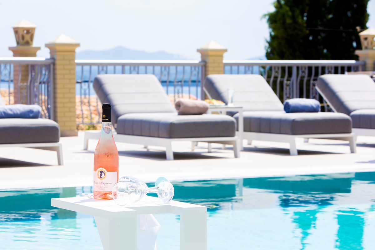 A sparkling pool glistens under bright sunlight, surrounded by several loungers in a neutral tone. A white side table holds a bottle of rosé and glasses, inviting relaxation. Lush greenery and distant mountains can be seen in the background.