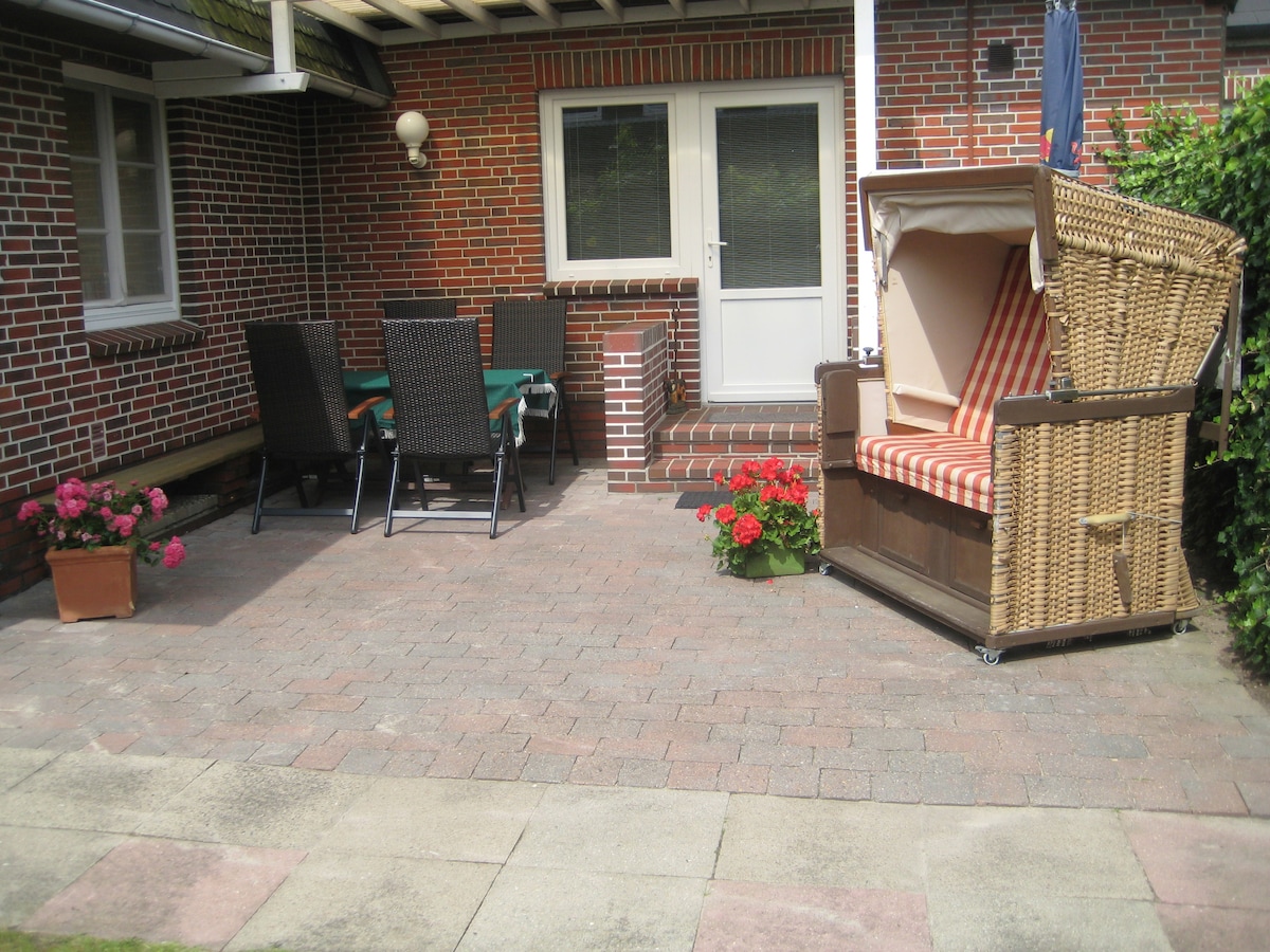 A private terrace is shown, featuring a dining set with four black chairs and a green table. A traditional beach chair is positioned beside vibrant flower pots with red blooms. The area is paved and surrounded by a brick wall, with a door leading into the apartment.