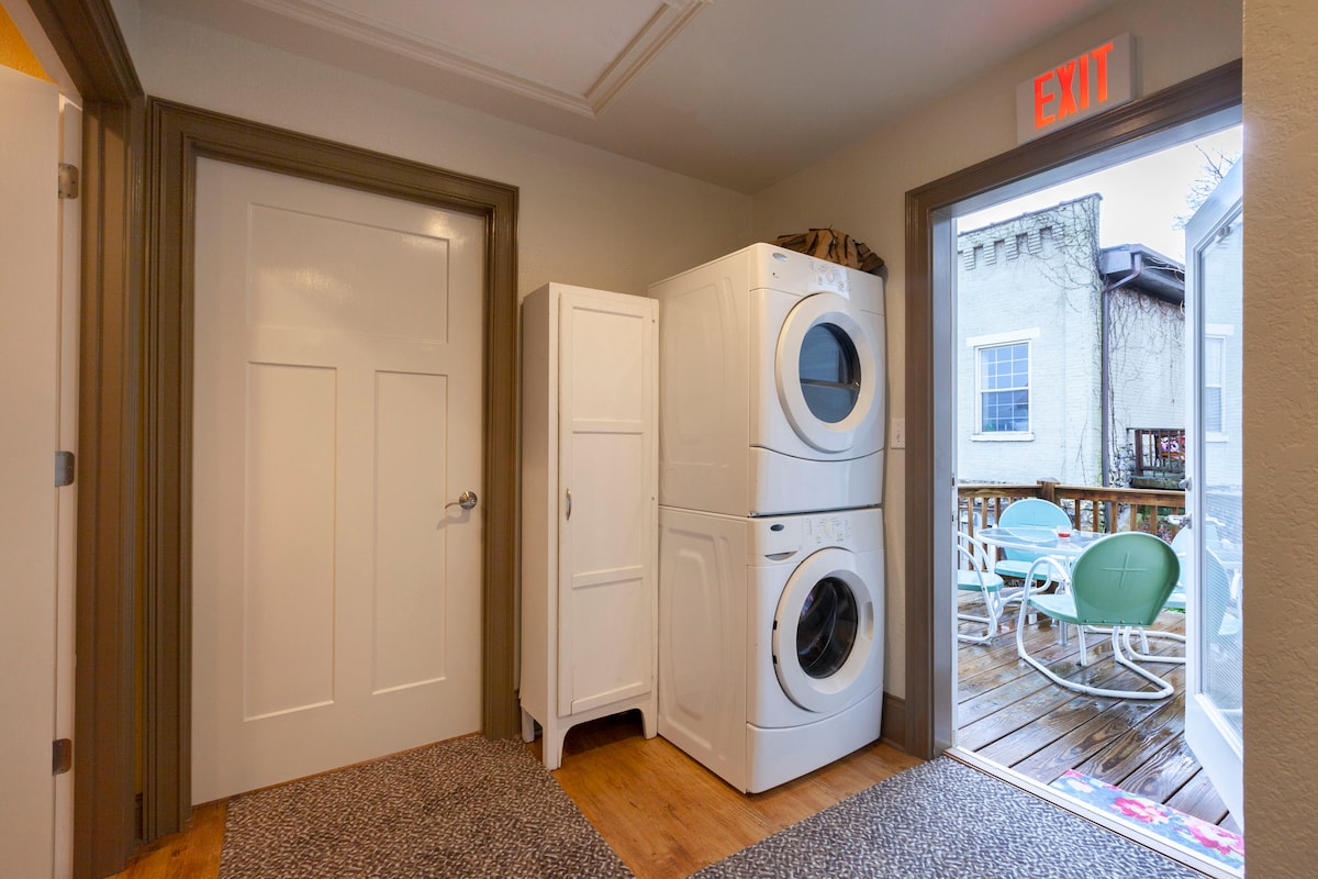 A functional laundry area is visible, featuring stacked appliances beside a white cabinet. A door leads to an outdoor deck with seating. Natural light filters in through a nearby window, complementing the neutral tones of the space.