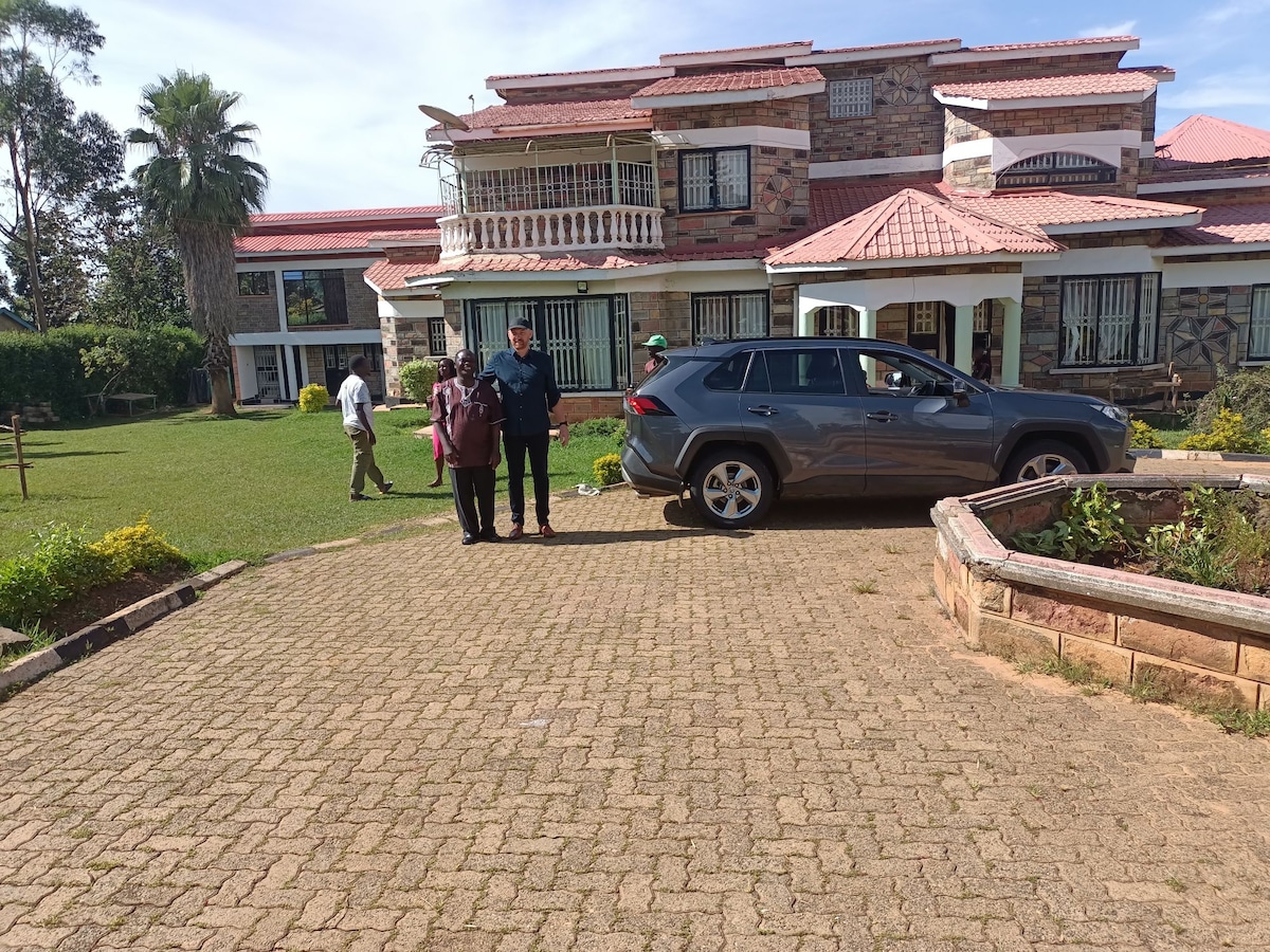 A spacious guesthouse is showcased, featuring a façade of stone and brick with a red-tiled roof. In the foreground, a well-maintained garden surrounds a gravel driveway, where two individuals stand near a parked gray SUV. Lush greenery and a serene environment are visible in the background.