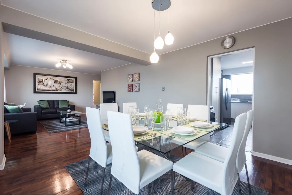 A spacious dining area is illuminated by pendant lights, featuring a large glass table surrounded by white chairs. A cozy living room can be seen in the background, with a dark sofa and a decorative area rug.