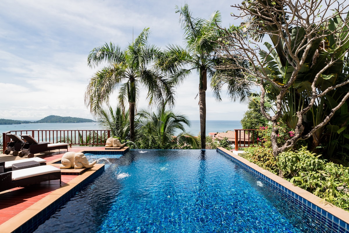 A tranquil infinity pool stretches towards the horizon, bordered by lush tropical foliage. Comfortable loungers are positioned on a wooden deck, inviting relaxation. In the background, panoramic views of the azure sea are visible, under a bright sky dotted with soft clouds.