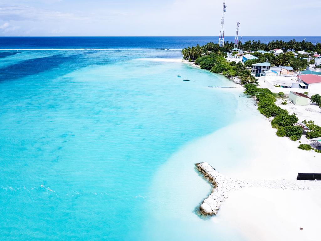 An aerial view captures the turquoise waters gently meeting the white sandy shoreline. Lush greenery and quaint structures dot the landscape, while rocky formations extend into the water, creating natural pathways. The vibrant blue skies enhance the serene coastal scene.