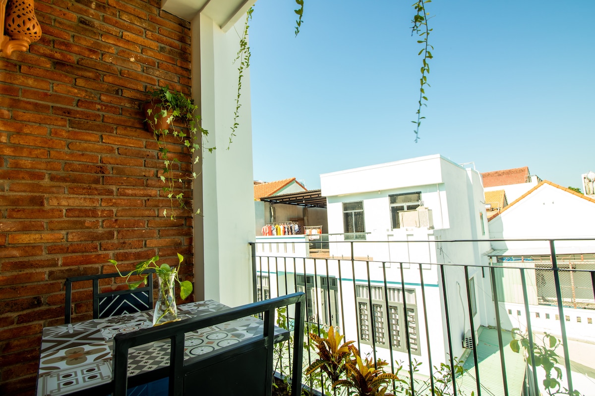 A balcony area features a table set with chairs, surrounded by climbing plants against a textured brick wall. The view includes nearby buildings under a clear blue sky, creating an inviting outdoor space for relaxation.