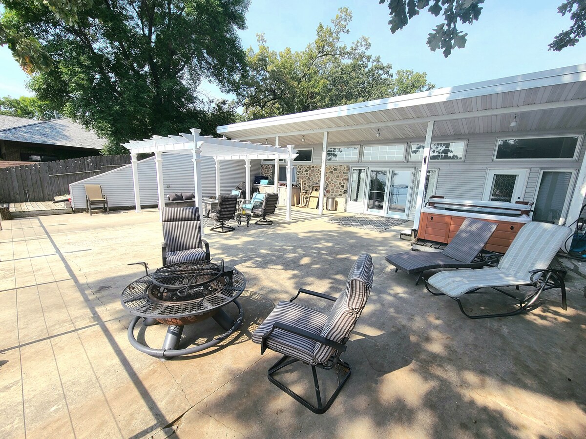 A spacious outdoor patio is shown, featuring several lounge chairs and a fire pit surrounded by trees. A covered area houses a hot tub, and large glass doors open into the interior of the home, enhancing connectivity between indoor and outdoor spaces.