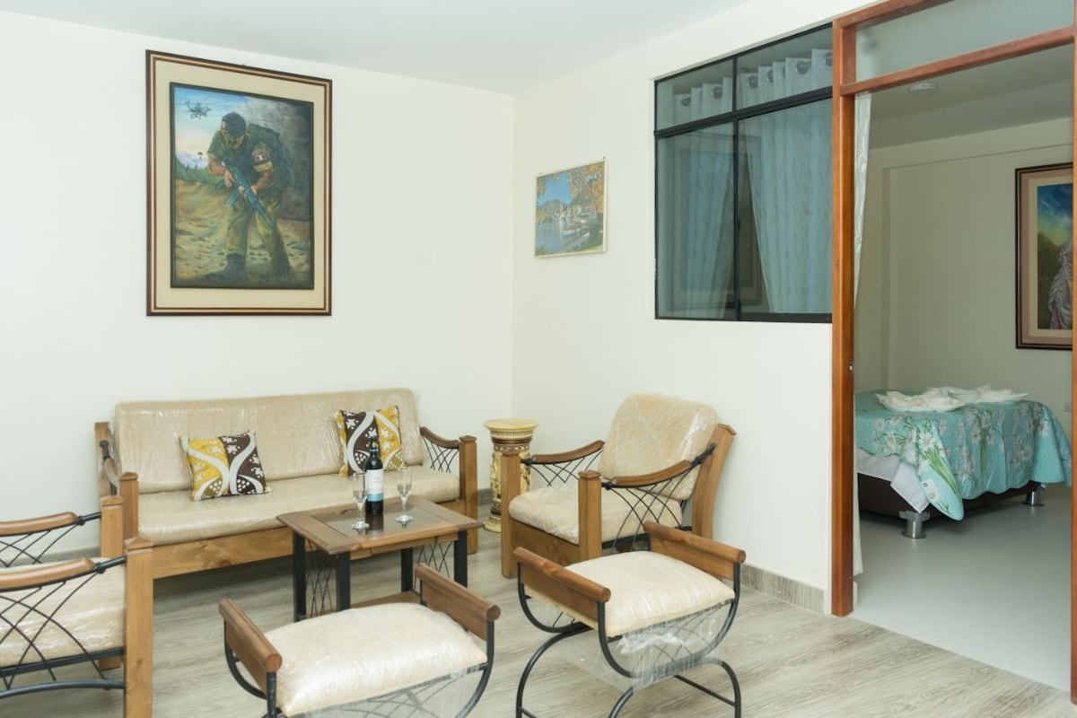 A living area features a light-colored sofa and wooden chairs with decorative cushions, arranged around a small wooden table. Artwork adorns the walls, and large windows allow natural light to fill the space. A glimpse of a bedroom is visible through an open doorway.