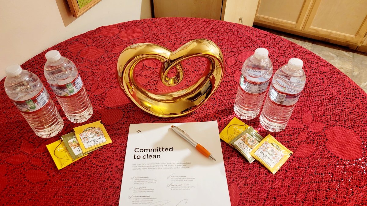 A round table is covered with a textured red tablecloth featuring a decorative golden heart sculpture at its center. Three bottles of water are arranged symmetrically alongside the sculpture, with packets of tea situated nearby. A document labeled 'Committed to clean' and a pen are placed on the table.