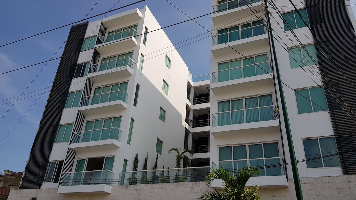 A modern residential building is depicted from the outside, featuring multiple floors with large glass windows. The façade presents a mix of white and dark tones, framed by green plants at the base. Clear blue skies provide a bright backdrop to the structure.