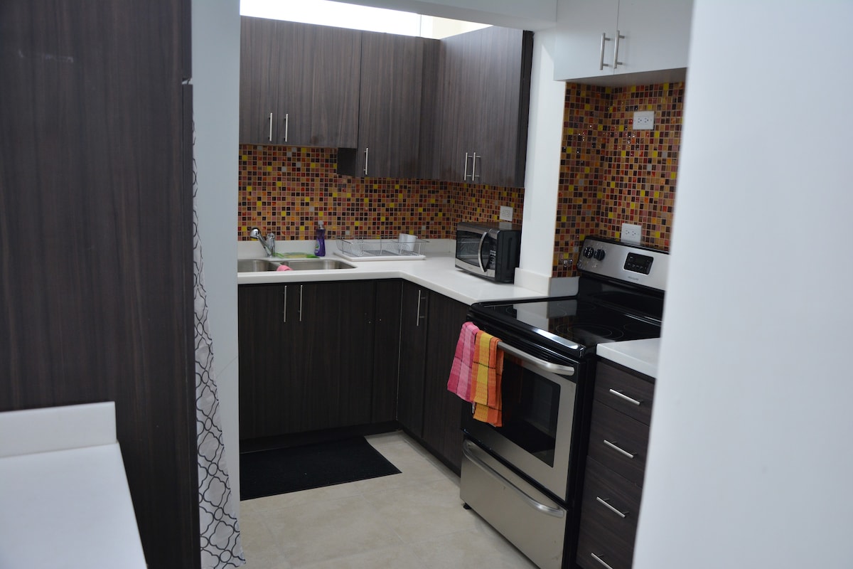 The kitchen features modern cabinetry with a mosaic tile backsplash in warm colors. A stainless steel stove and microwave are positioned alongside a countertop with a double sink. A colorful towel hangs from the oven handle, and the floor is finished with smooth tiles.