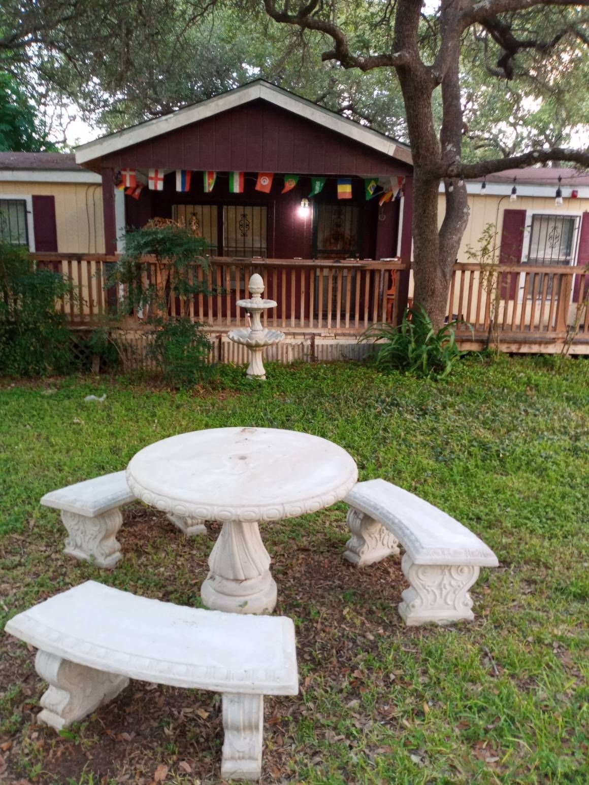 A stone table surrounded by four matching benches occupies a grassy area, offering a space for outdoor relaxation. The house in the background features a porch with colorful flags and is framed by trees, enhancing the peaceful setting.