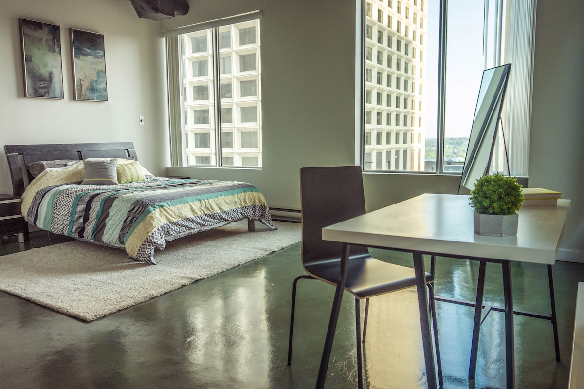 A spacious sleeping area features a bed with colorful bedding and decorative pillows. Natural light floods through large windows, highlighting a minimalistic desk with a small plant. A soft rug is placed underfoot, enhancing the comfort of the space.