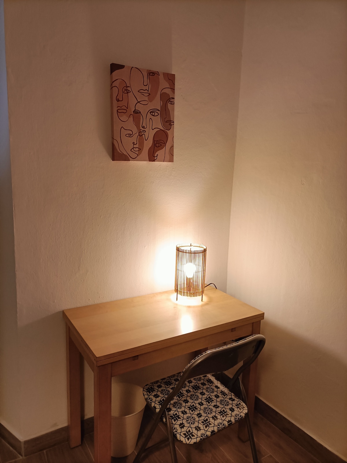 A small desk is positioned in a cozy corner, featuring a decorative lamp that provides soft lighting. Above the desk, an abstract artwork depicting stylized faces is mounted on the wall. A patterned chair is placed in front, creating an inviting workspace.