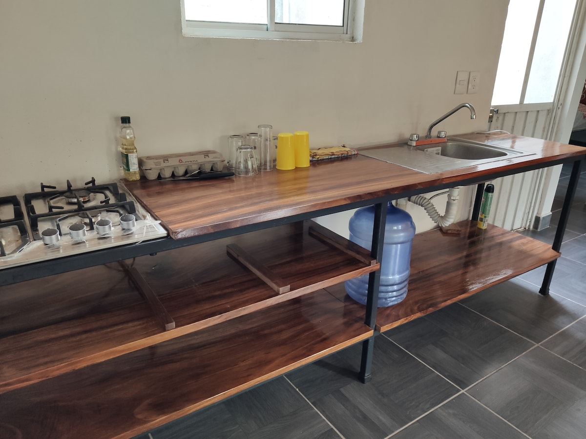 A functional kitchen area features a wooden countertop with a sink, a gas stove, and storage shelves below. A water dispenser is prominently placed, alongside a container of eggs and several glasses. Light enters from a nearby window, enhancing the workspace.