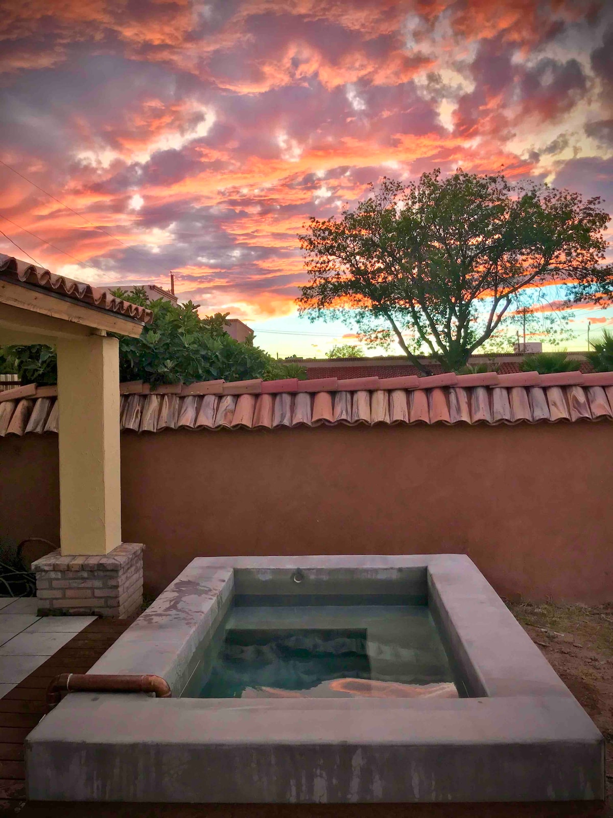A private hot mineral water bath is set against a colorful sunset, with hues of orange and pink filling the sky. The surrounding area features a garden setting with a tree and a terracotta-tiled roof structure, creating a serene escape.