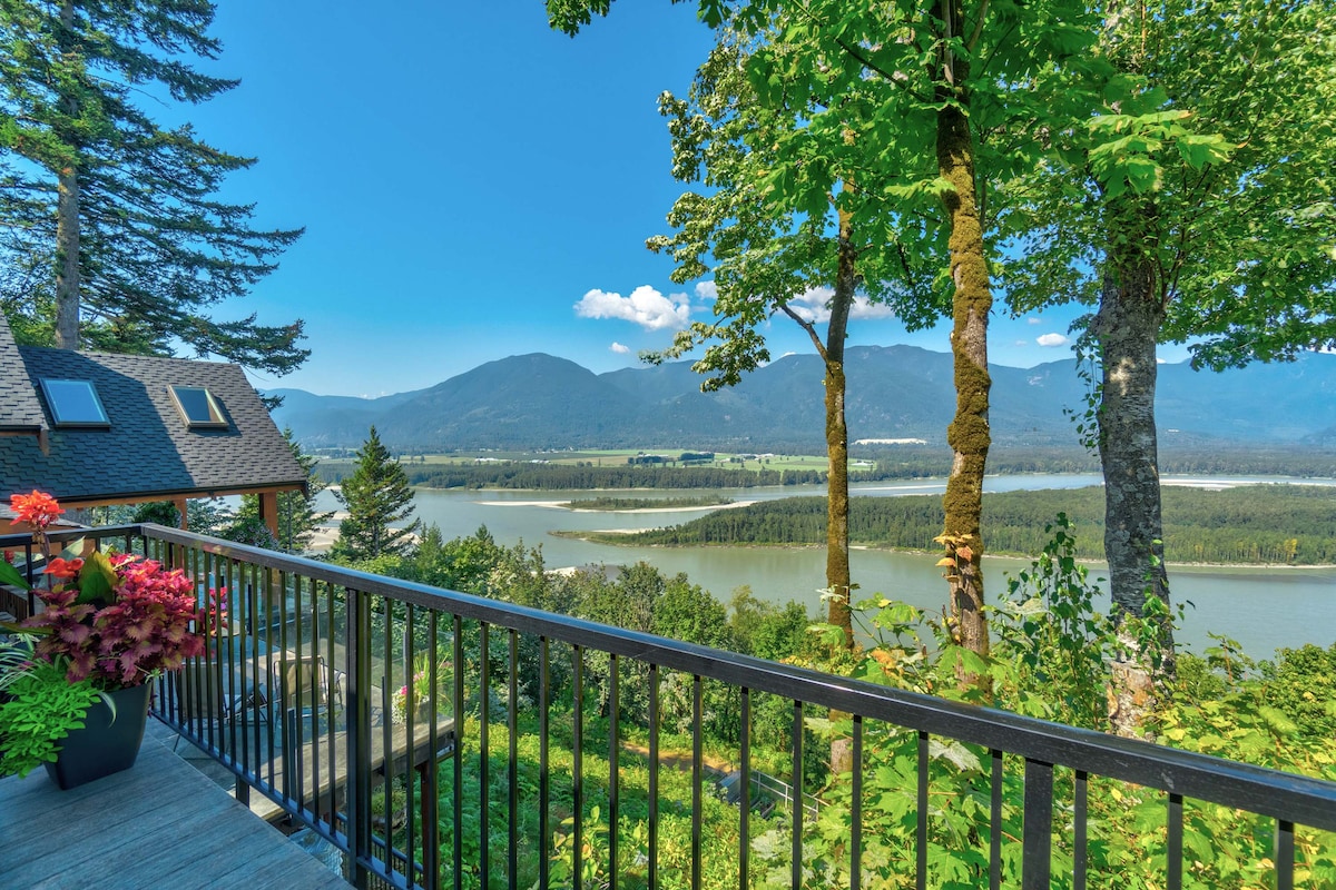 A scenic view from a deck overlooks the Fraser River and surrounding mountains. Lush green trees frame the image, while the sky displays a clear blue hue. Vibrant plants are positioned on the deck, enhancing the natural beauty of the outdoor space.